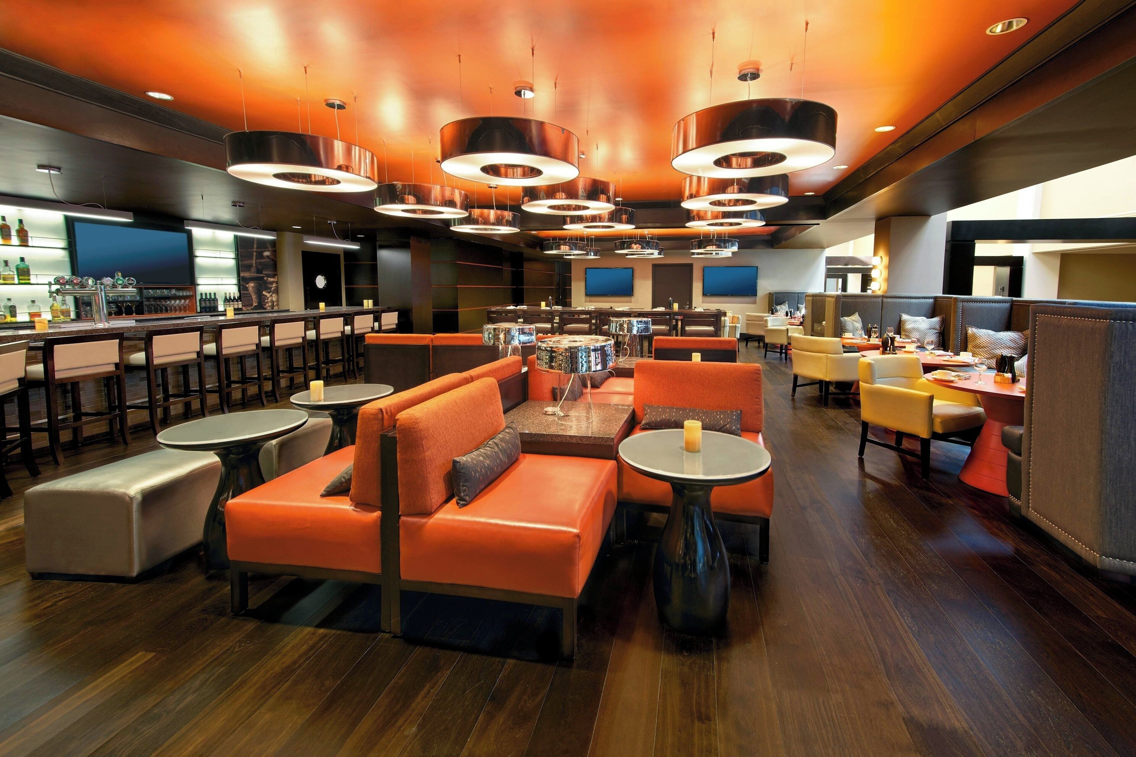 A modern bar area with orange seating, a bar counter, and overhead circular lighting fixtures.