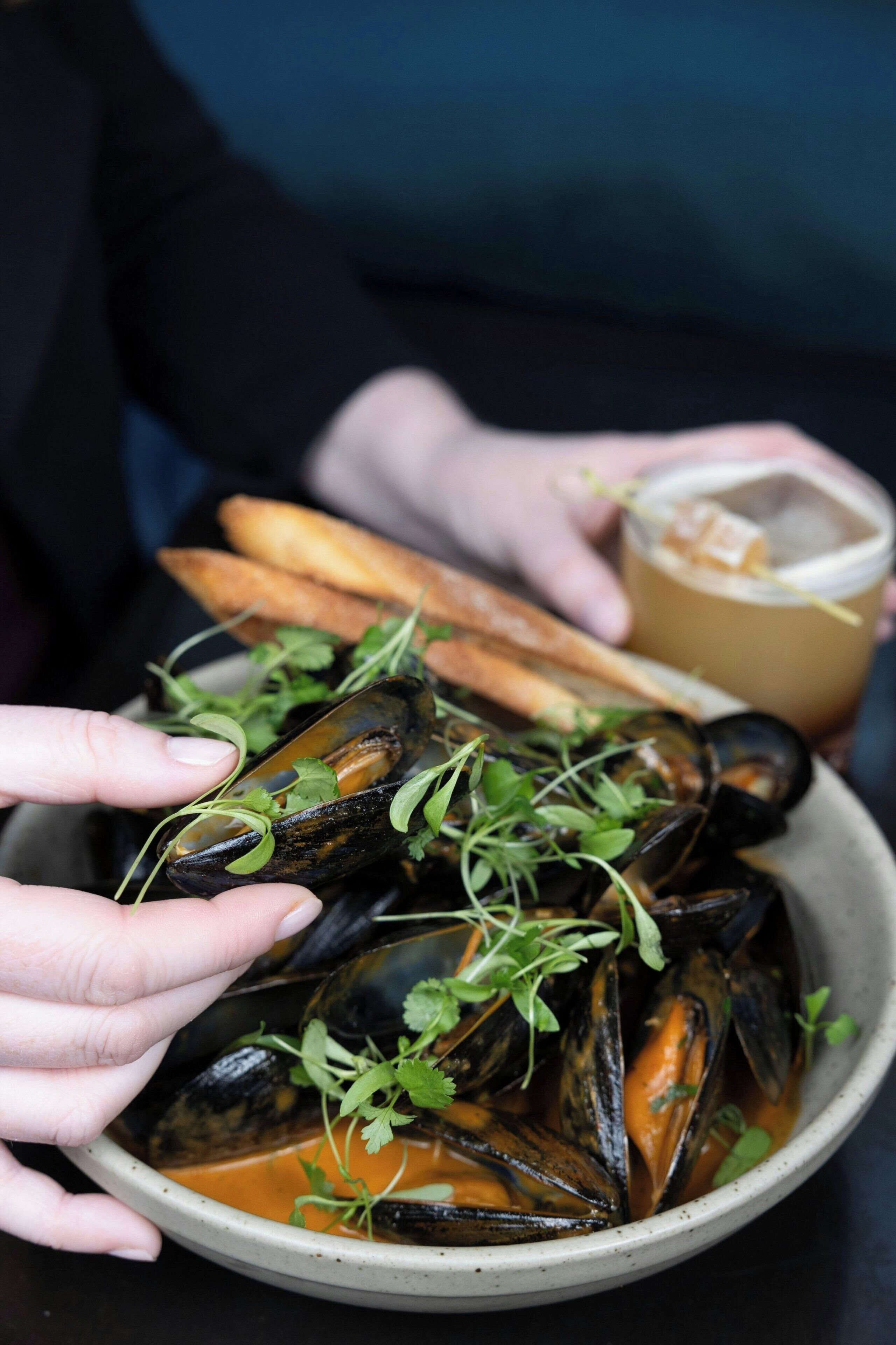 Una ciotola di cozze guarnite con erbe aromatiche, accompagnate da pane e una bevanda.