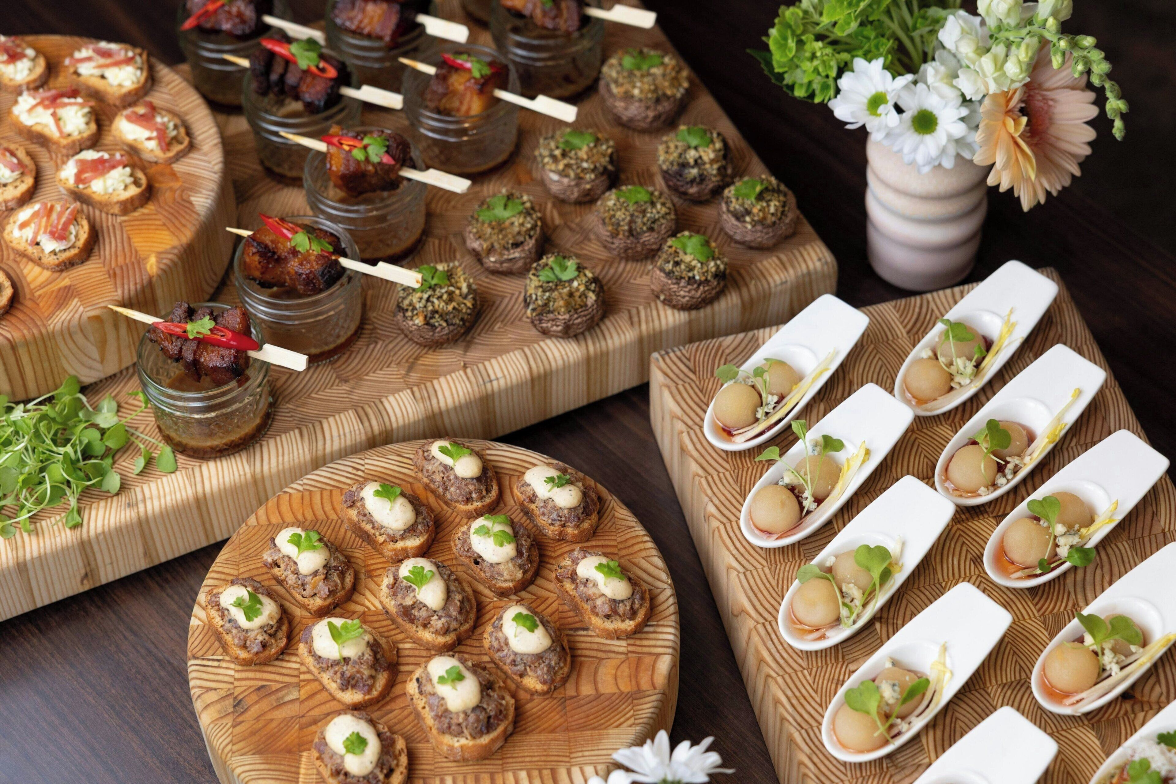 Diversos aperitivos dispuestos sobre tablas de madera y en frascos de vidrio, con un jarrón de flores al fondo.