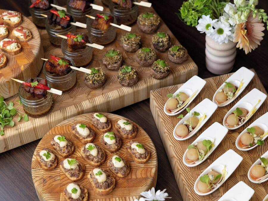 Diversos aperitivos dispuestos sobre tablas de madera y en frascos de vidrio, con un jarrón de flores al fondo.