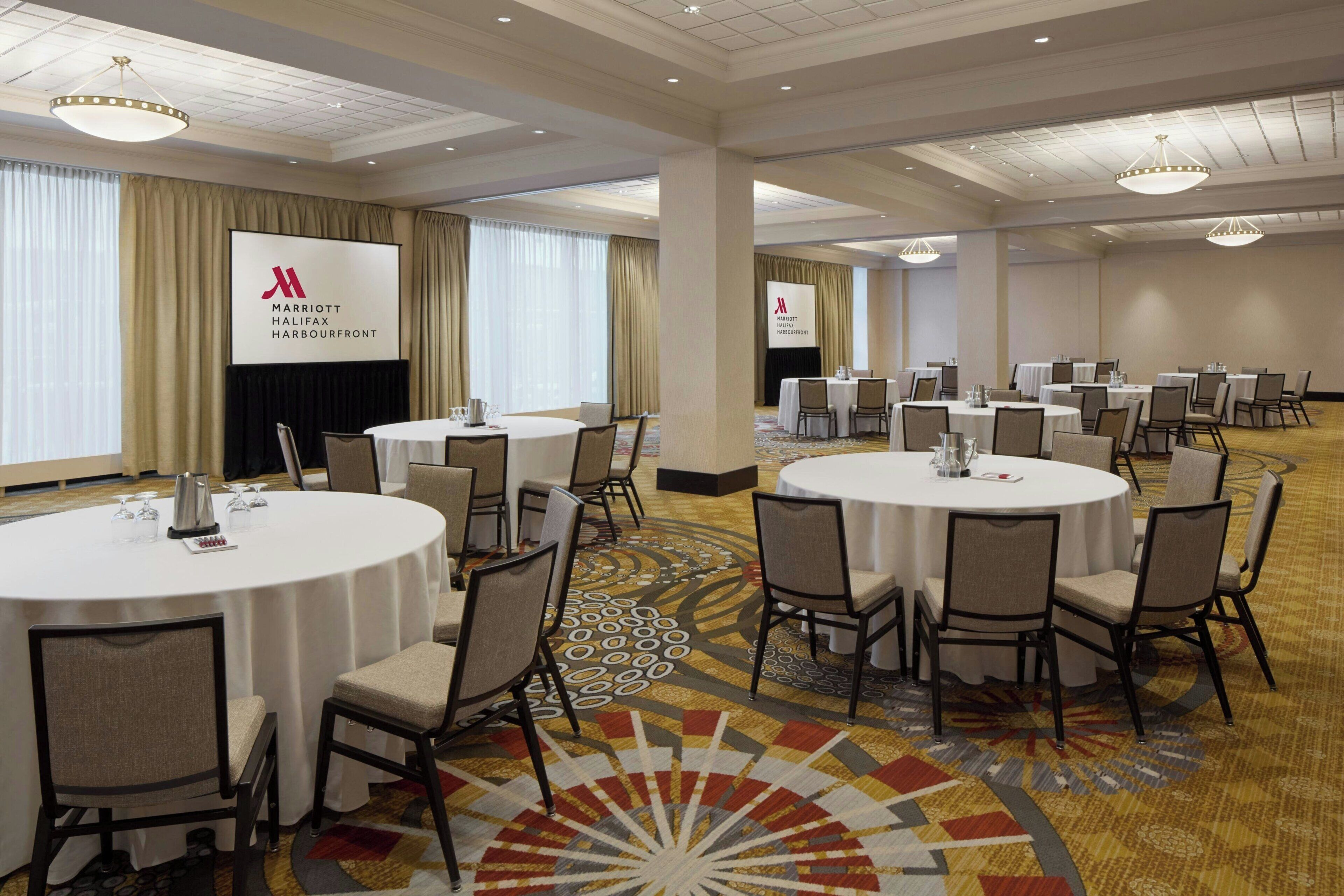 A hotel conference room with round tables, chairs, and a large screen displaying the hotel's logo.
