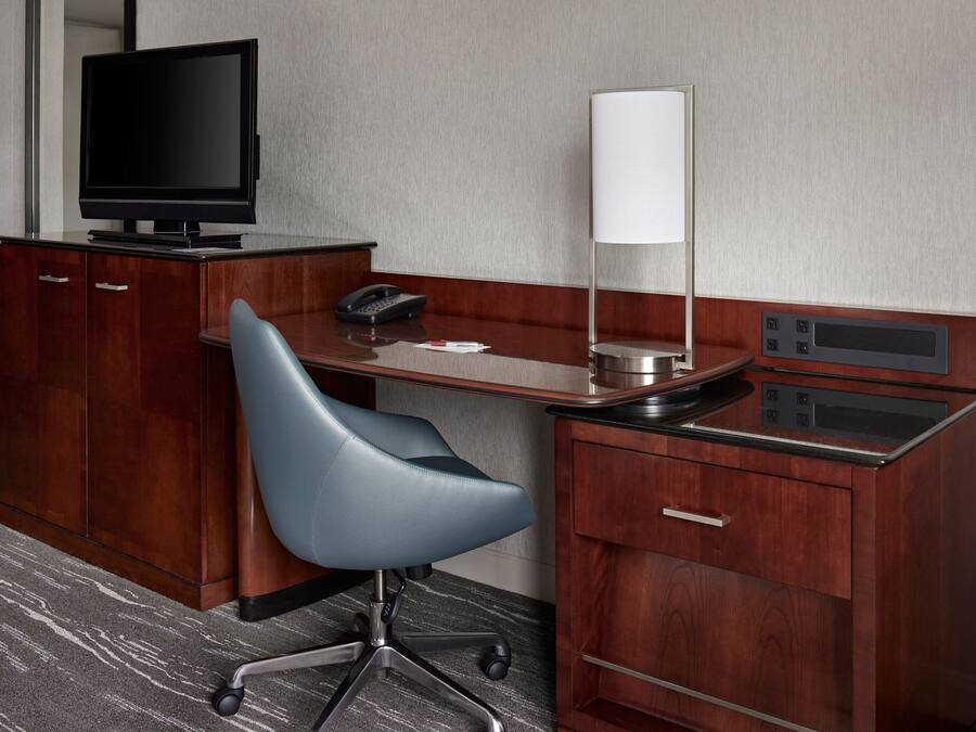Habitación de hotel con un escritorio de madera, una silla de oficina azul, un televisor de pantalla plana, un teléfono, una lámpara y una nevera minibar.