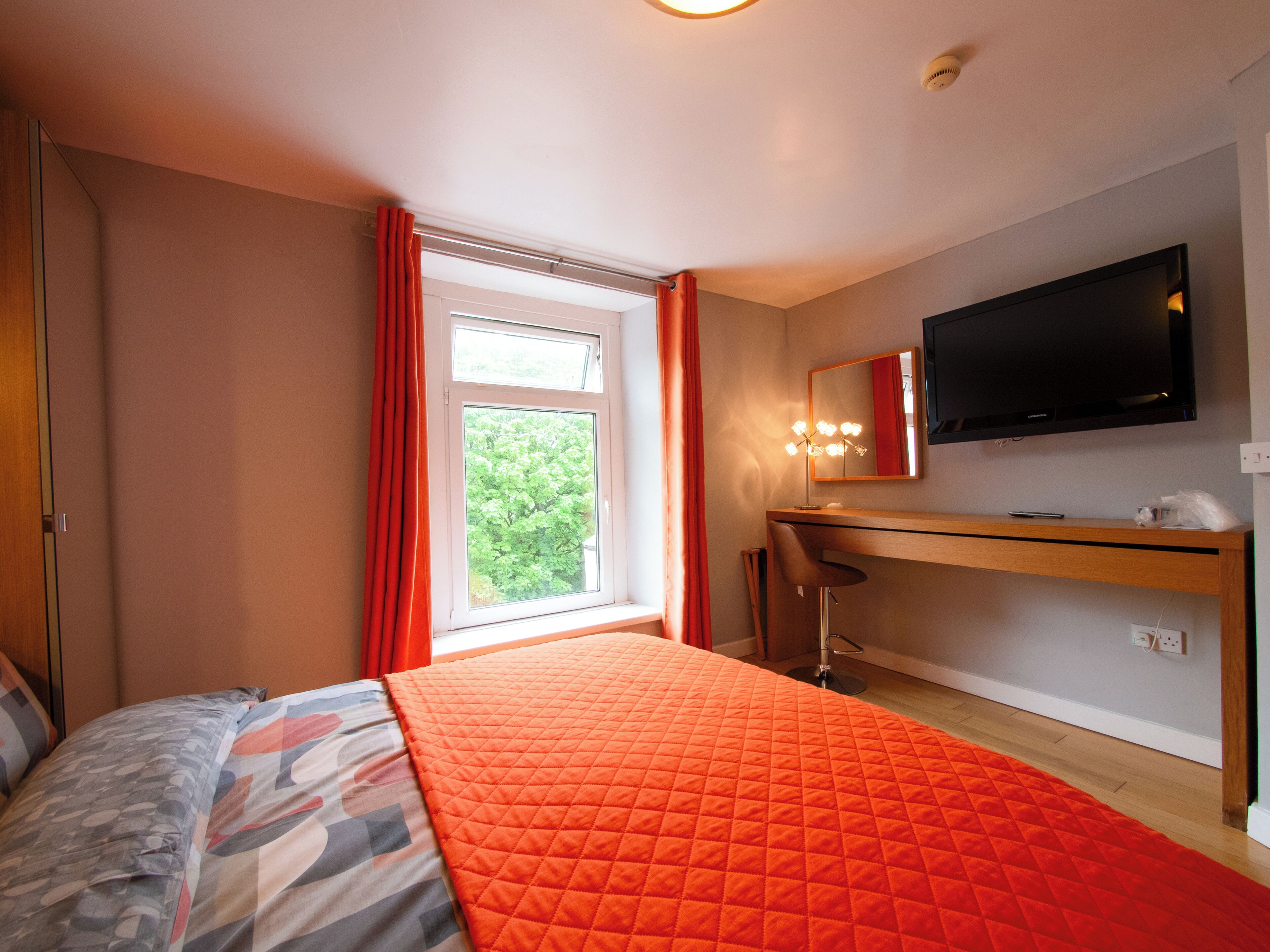 Une chambre à coucher comprenant un lit, un bureau, une chaise, un téléviseur et une fenêtre avec des rideaux.