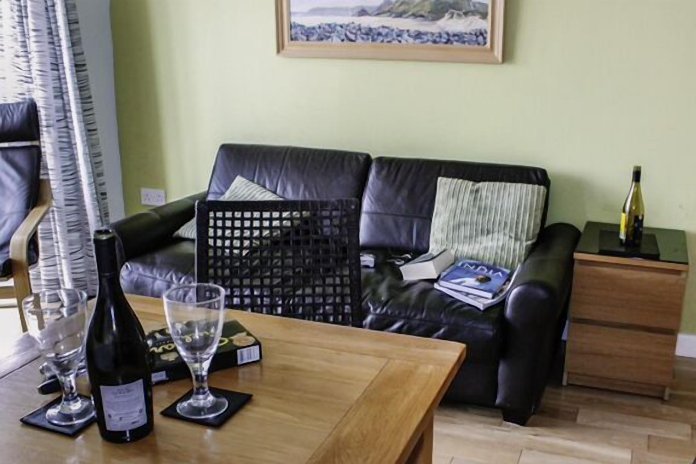 Uma sala de estar com um sofá de couro preto, uma mesa de centro de madeira com garrafas de vinho e copos, uma moldura com uma imagem na parede e uma pequena mesa lateral de madeira com uma garrafa.