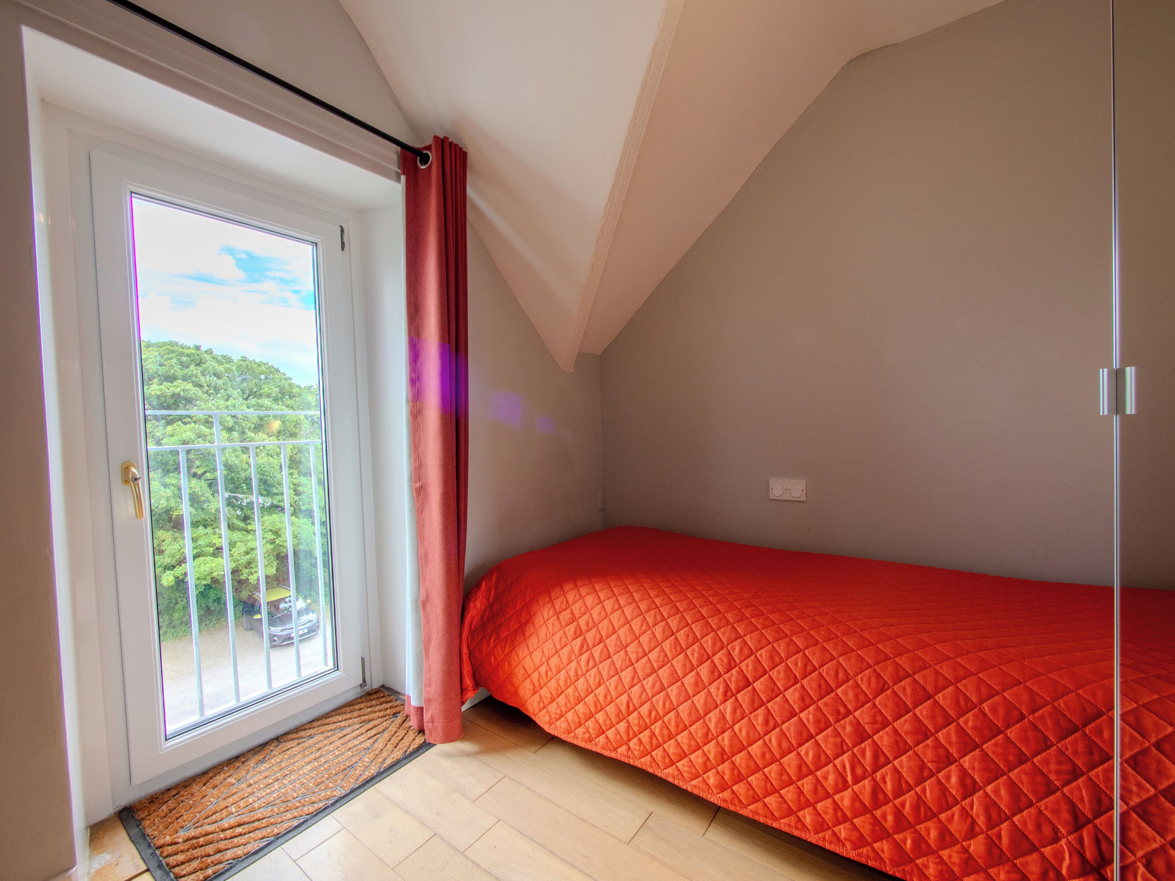 A small, single-bed room with a red quilted bedspread, a sliding glass door leading to a balcony, and a wooden floor.
