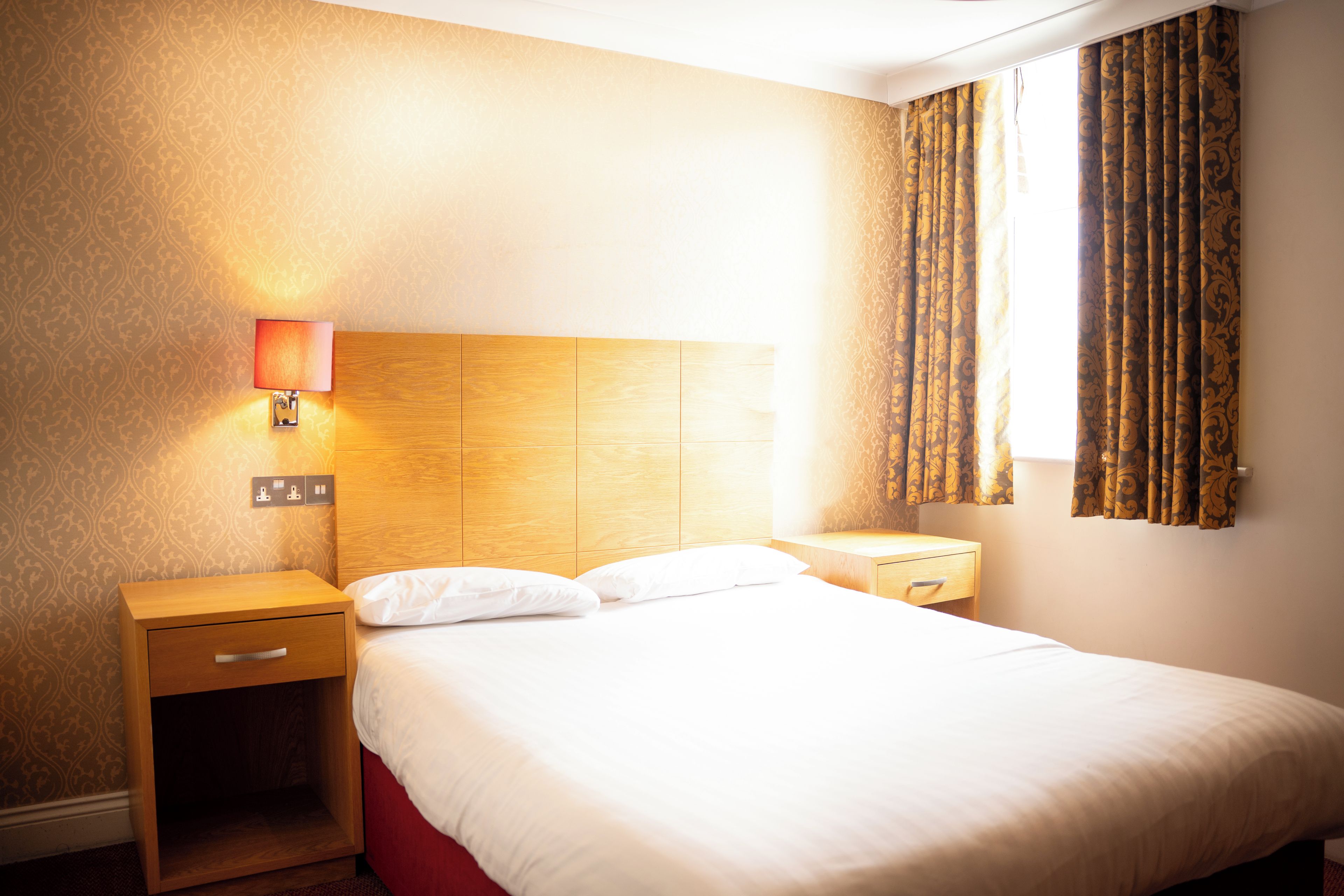 A hotel room with a bed, a nightstand, a lamp, and a window with curtains.