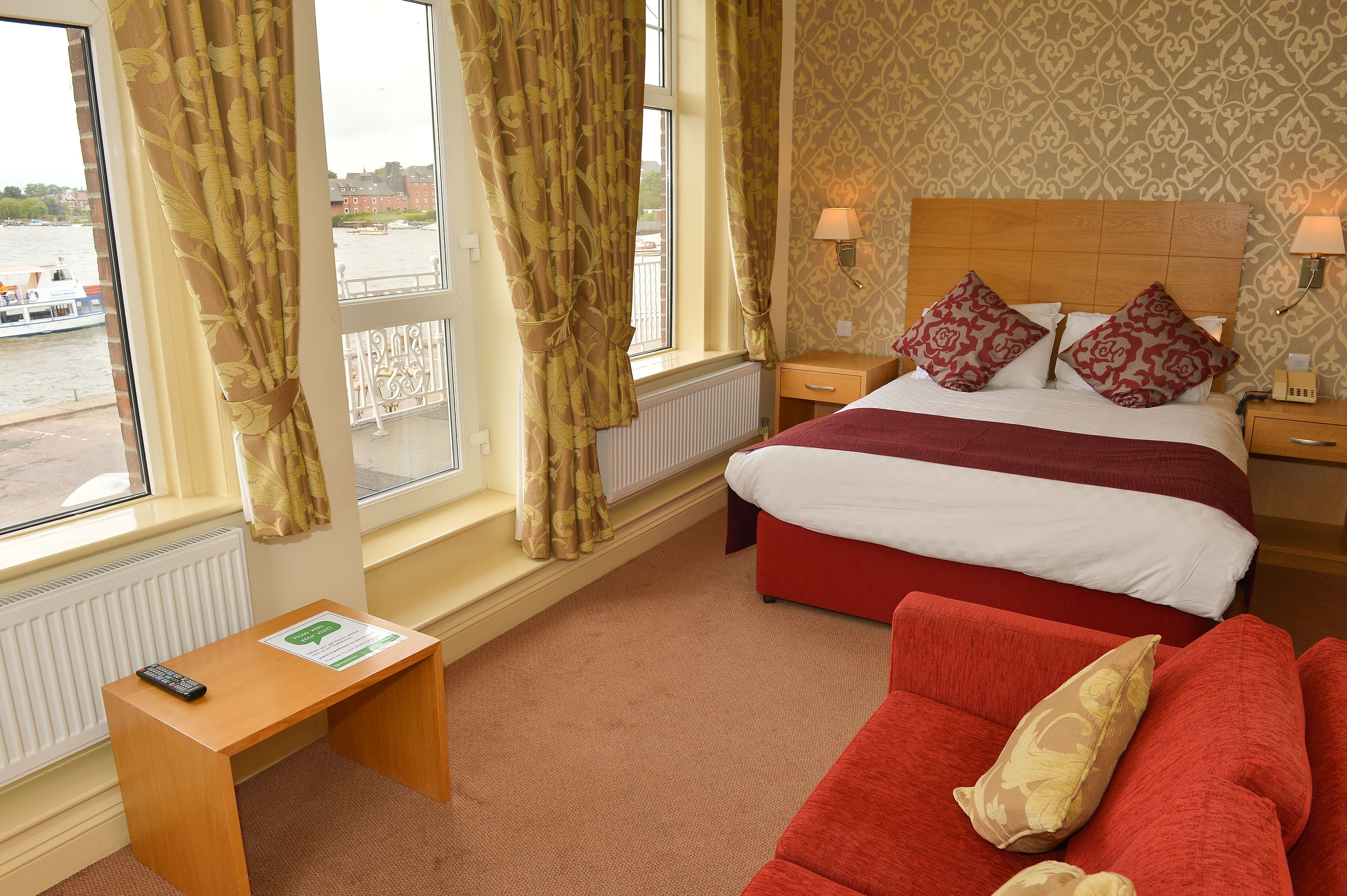 A hotel room with a bed, a red sofa, a wooden nightstand, a bedside table with a remote control, and a window with curtains.