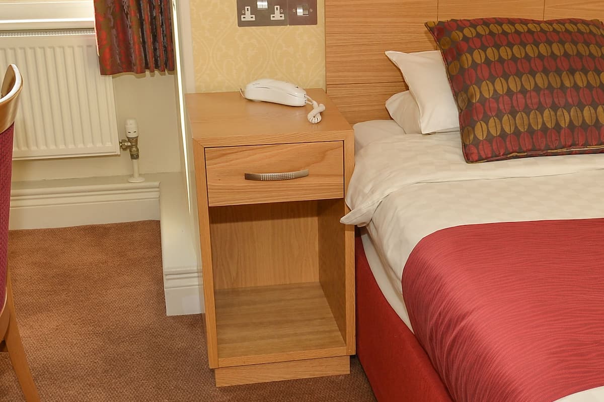 A hotel room with a wooden nightstand, a white telephone, a bed with a red and beige bedspread, and a patterned throw pillow.