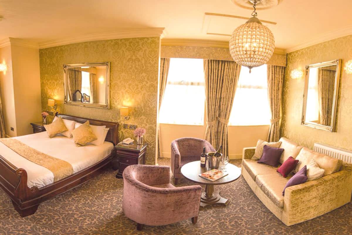 A hotel room with a large bed, a sitting area with a sofa, a desk, and a chair. There are two chandeliers, a mirror, and a window with curtains.