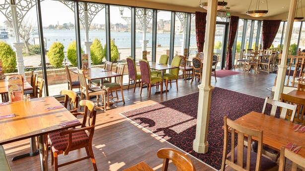 A restaurant with outdoor seating overlooking a body of water, featuring wooden tables and chairs, and a view of the water and buildings in the distance. - Wherry Hotel (Lowestoft)