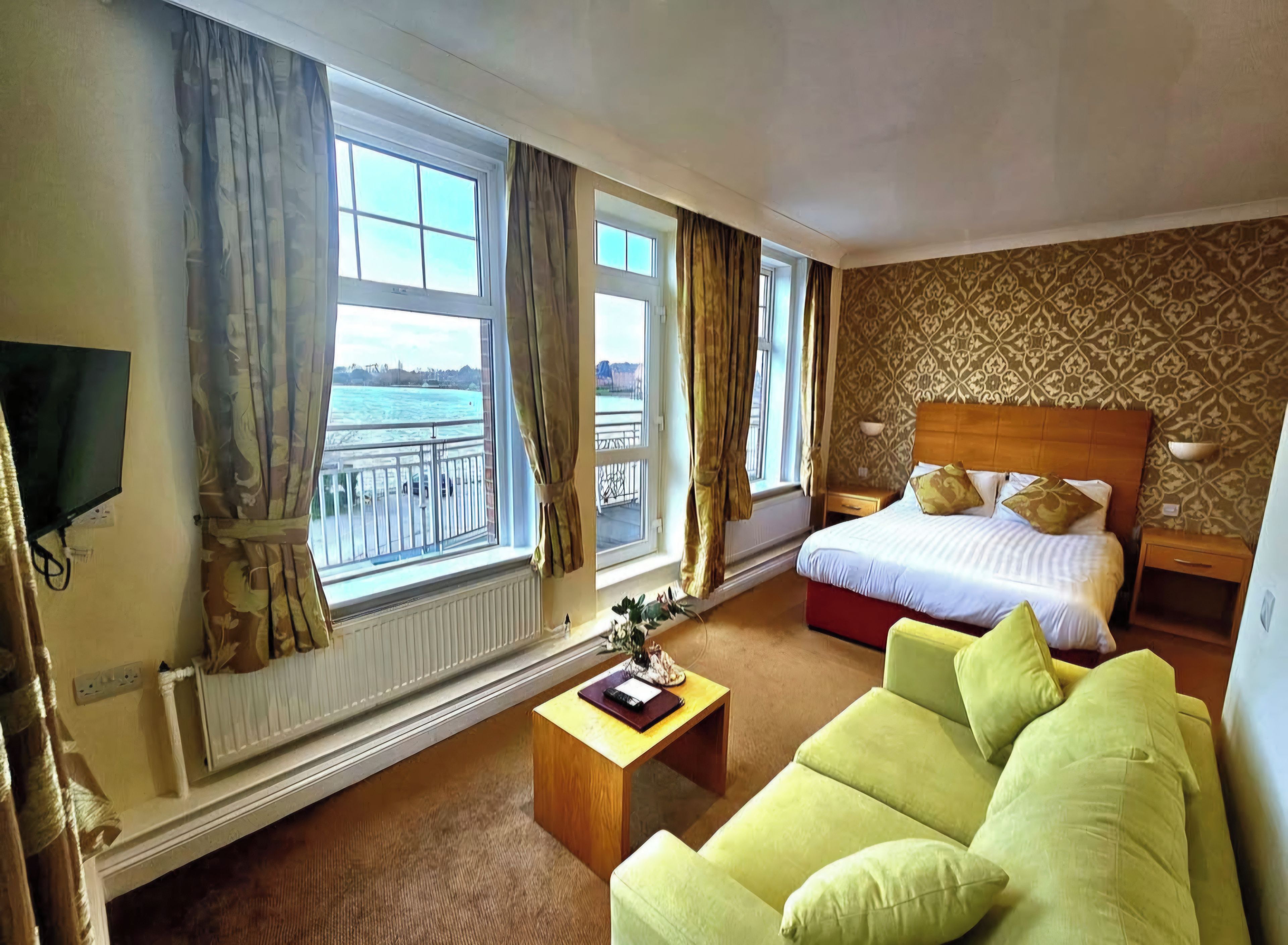 A hotel room with a bed, a green sofa, a wooden bedside table, a TV, and a window with curtains.