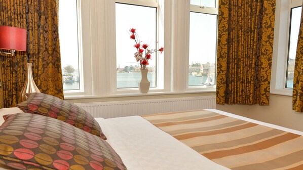 A hotel room with a bed, a vase with flowers, and a view of boats on the water through large windows.