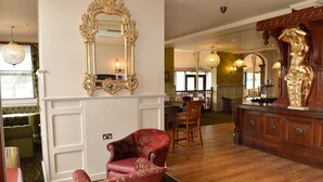 A spacious room with a large ornate mirror, a red upholstered chair, wooden flooring, and a bar area with a golden statue. - Wherry Hotel (Lowestoft)