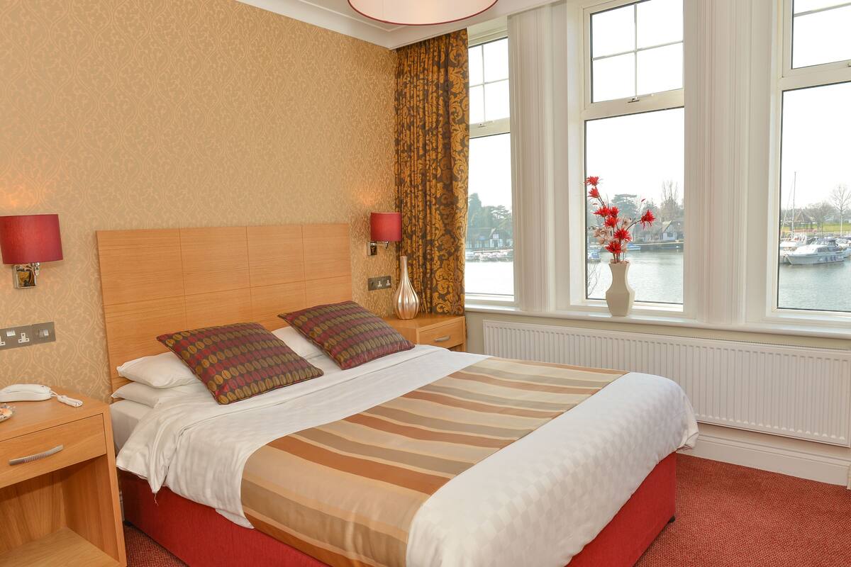 A hotel room with a large bed, two bedside lamps, a nightstand with a phone, and a view of boats through the window.