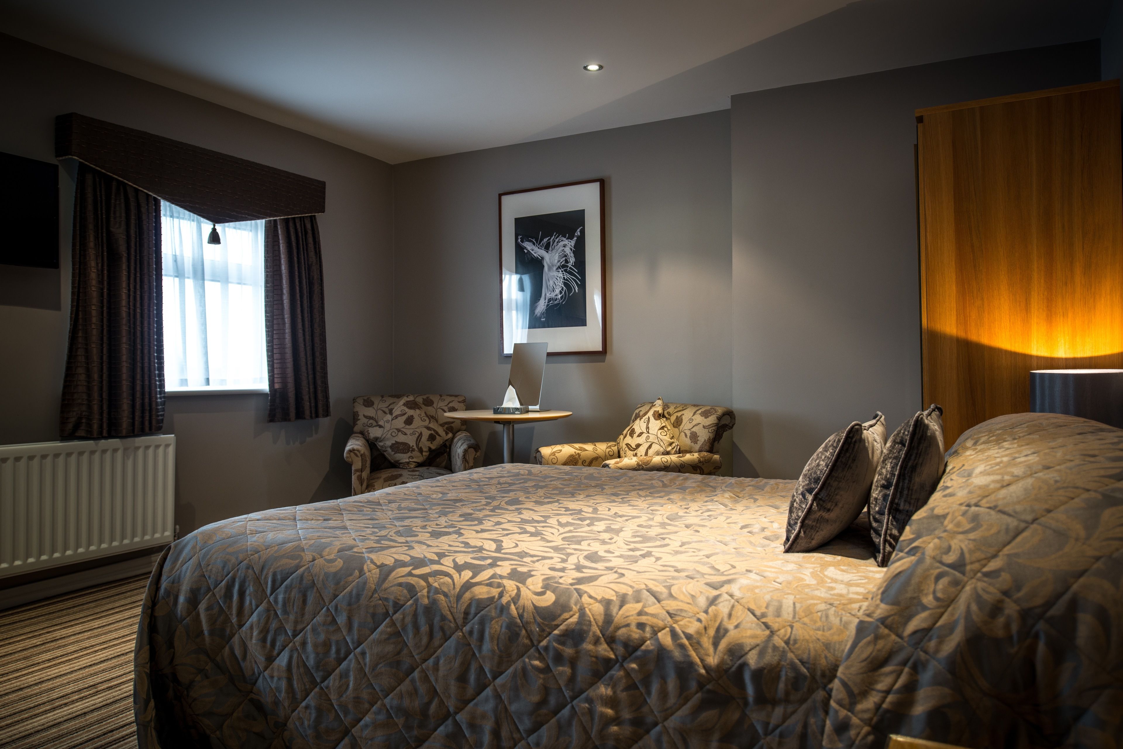 Habitación de hotel con una cama grande, una mesita, dos sillones, una obra de arte enmarcada en la pared y un ventanal con cortinas.