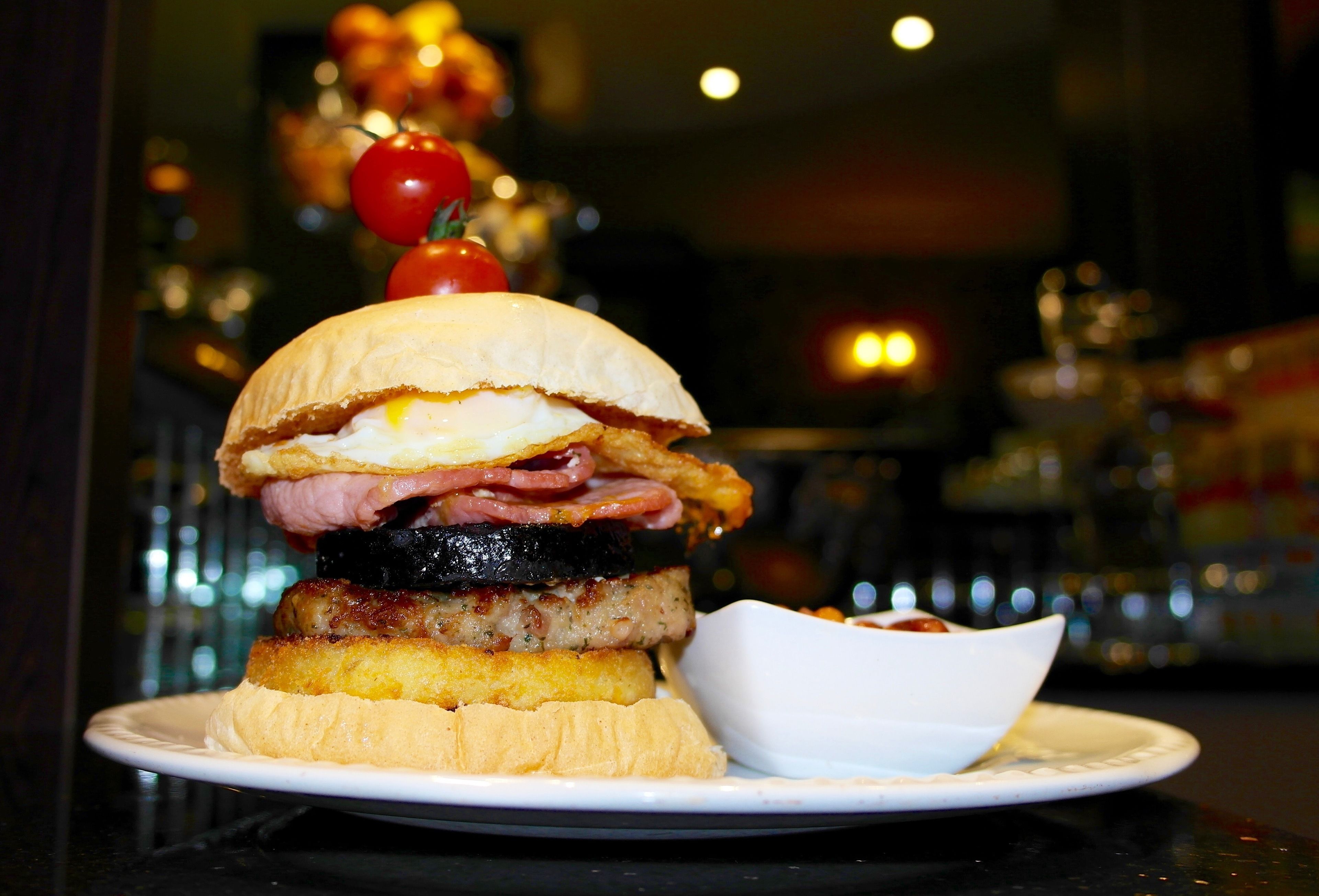 A sandwich with multiple layers of meat and egg on a bun, served on a plate with a small bowl of sauce.