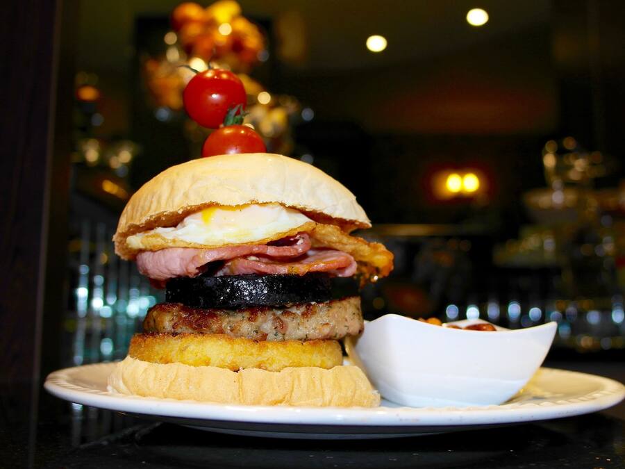 A sandwich with multiple layers of meat and egg on a bun, served on a plate with a small bowl of sauce.