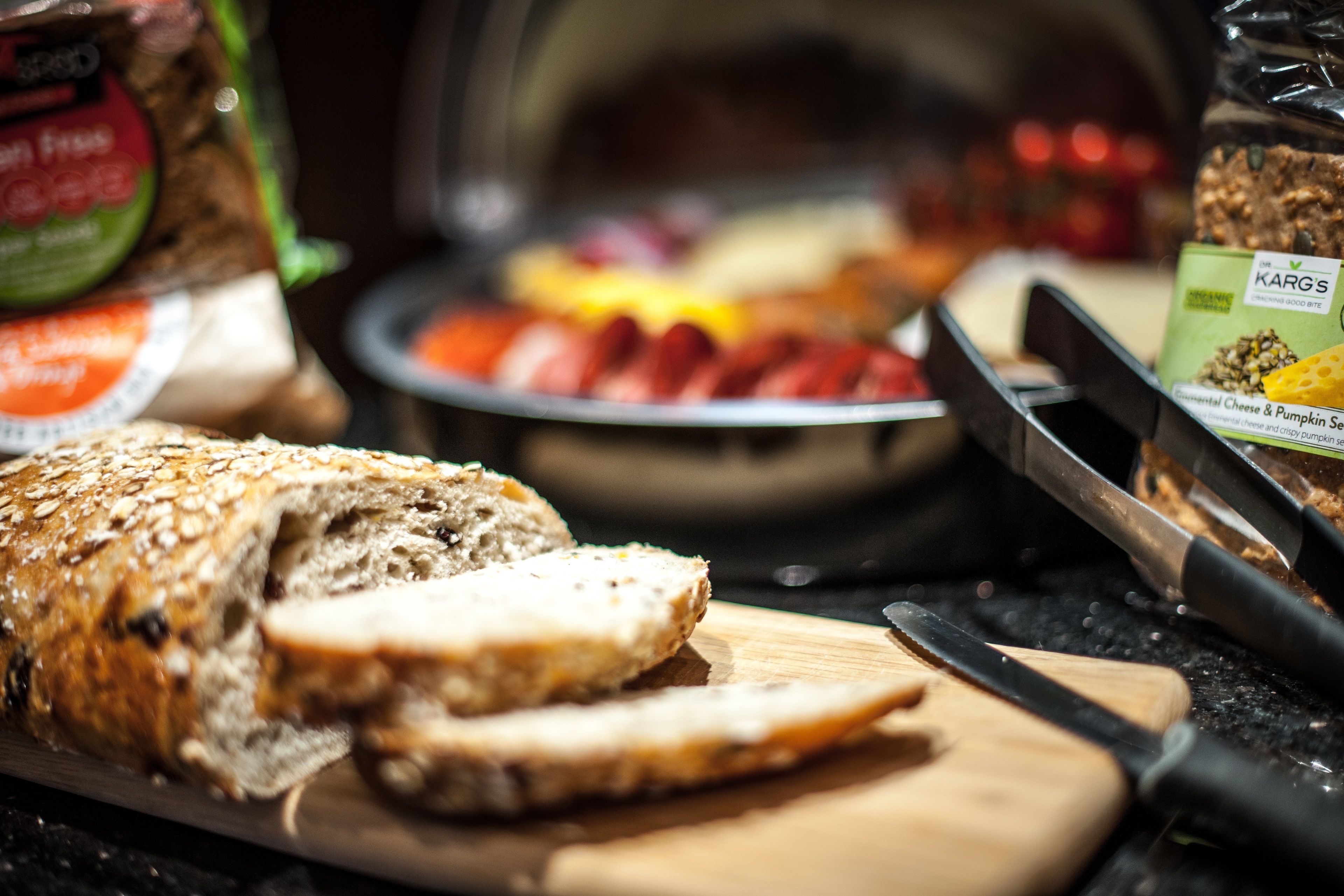 A loaf of sliced bread on a cutting board with a knife, a bowl of sliced tomatoes, and a jar of Karg's cheese & pumpkin seeds.