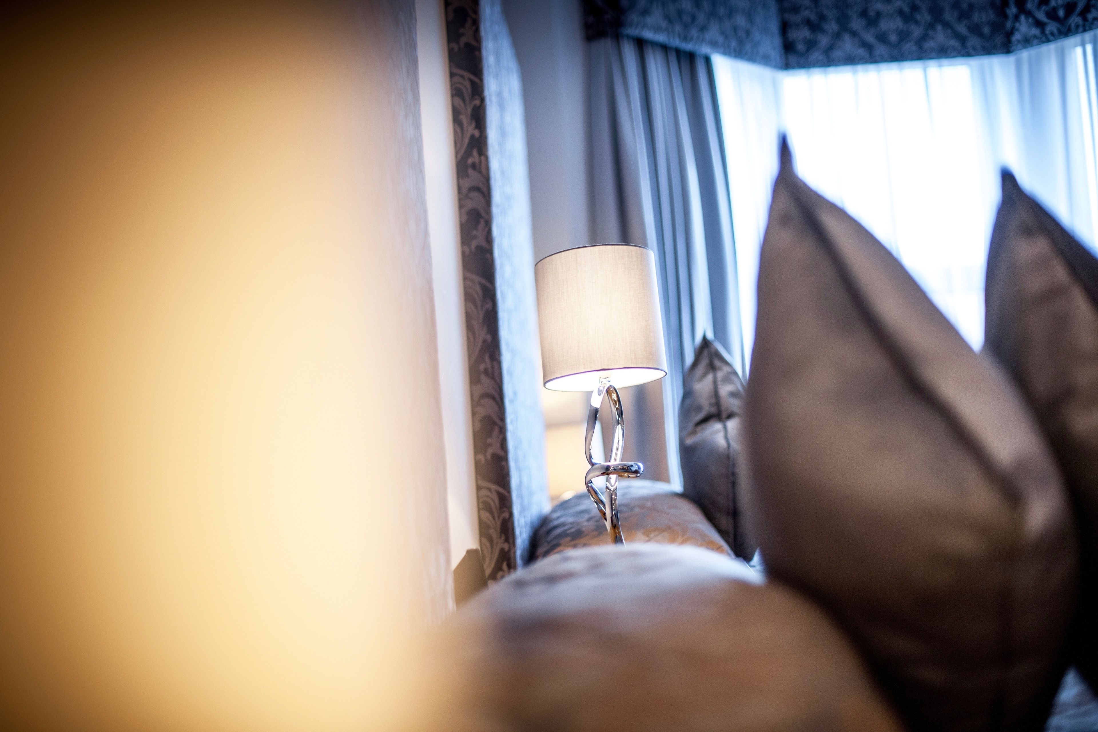 Een close-up van een lamp op een nachtkastje, met uitzicht op een raam met gordijnen en kussens.