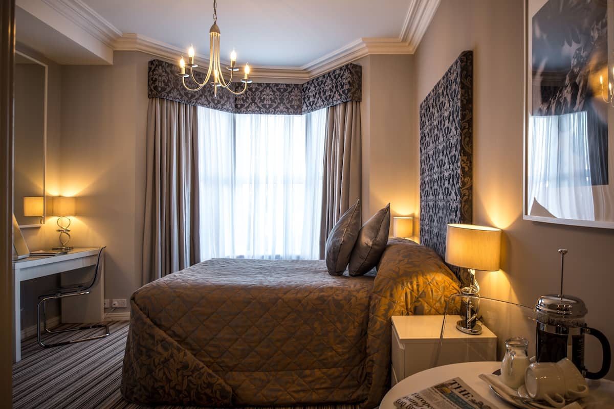 A hotel room with a large bed, a desk, a chair, a coffee machine, and a lamp.