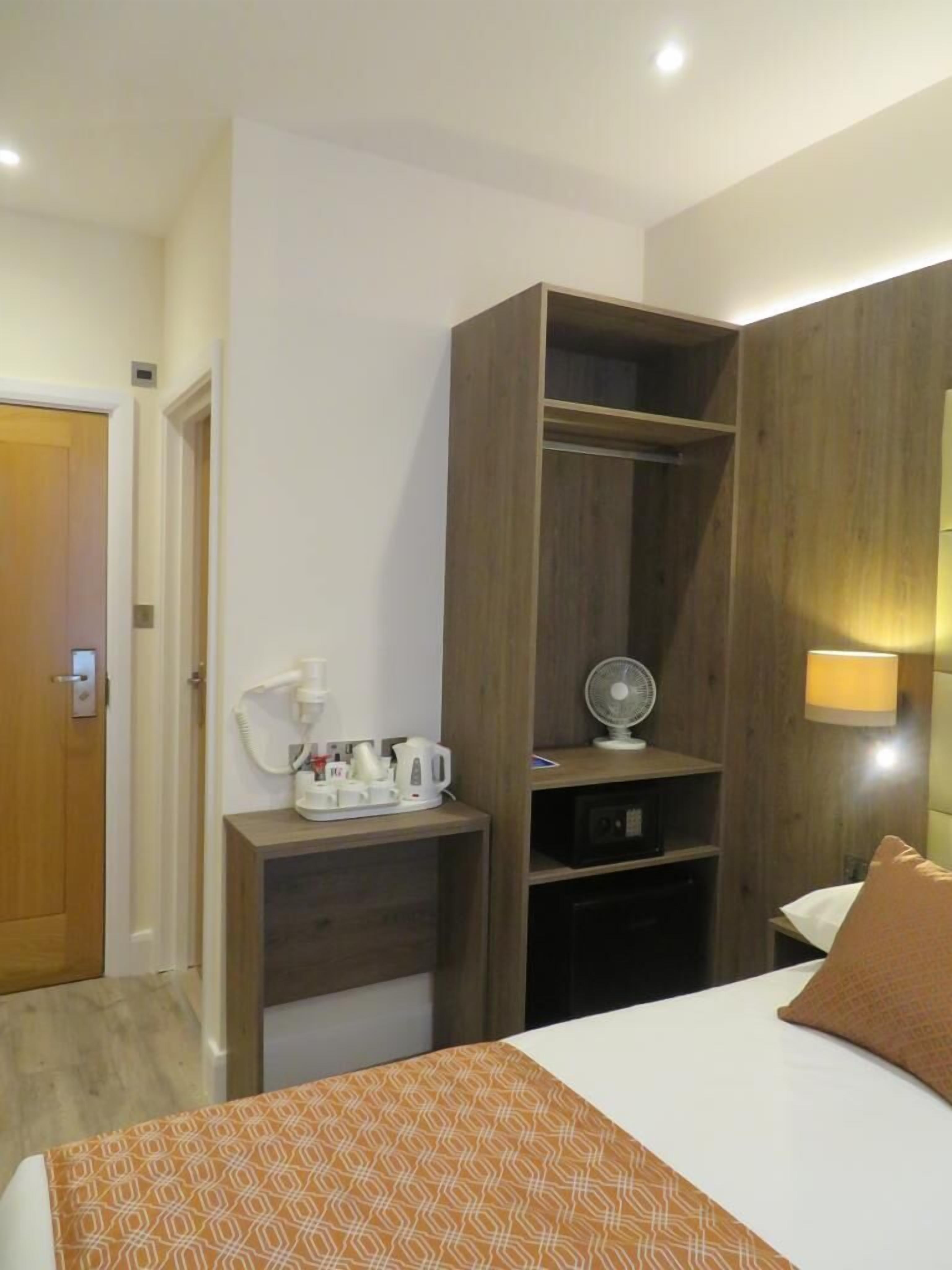 A hotel room with a bed, a bedside table with a lamp, a wooden wardrobe, and a small shelf with a fan and a kettle.