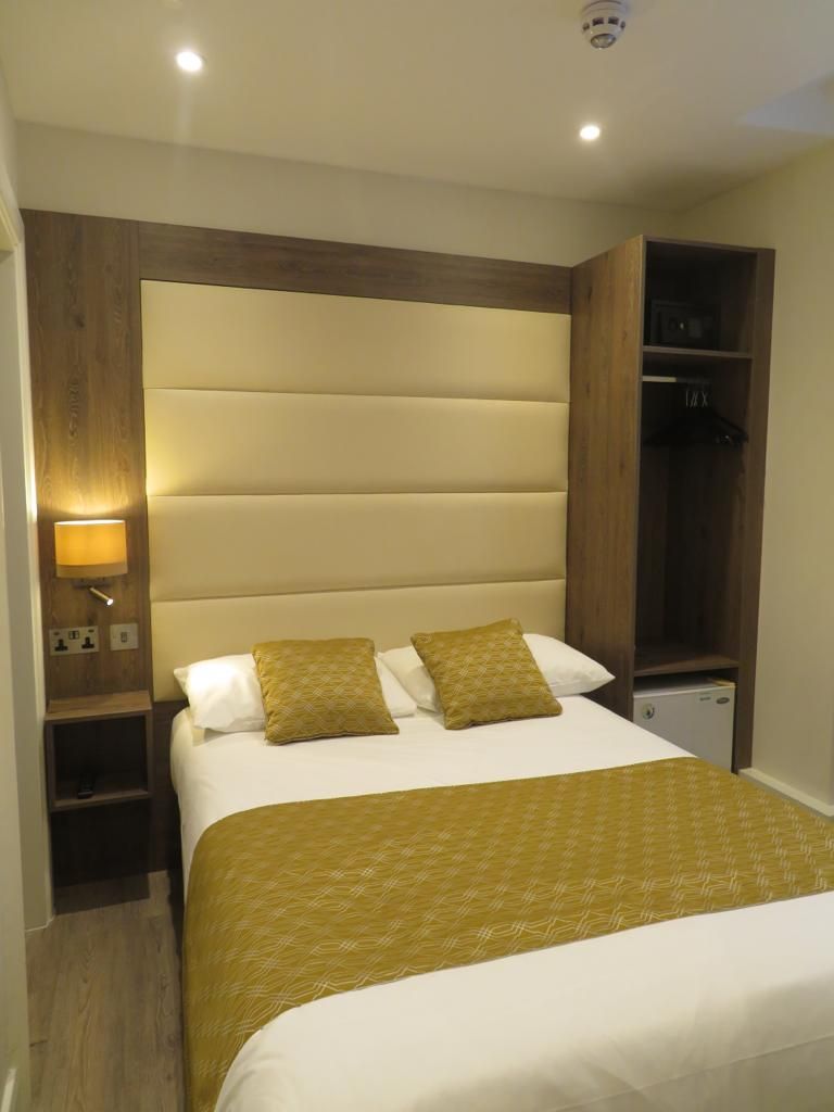 A hotel room with a large bed, a wooden headboard, a bedside table with a lamp, and a wardrobe.