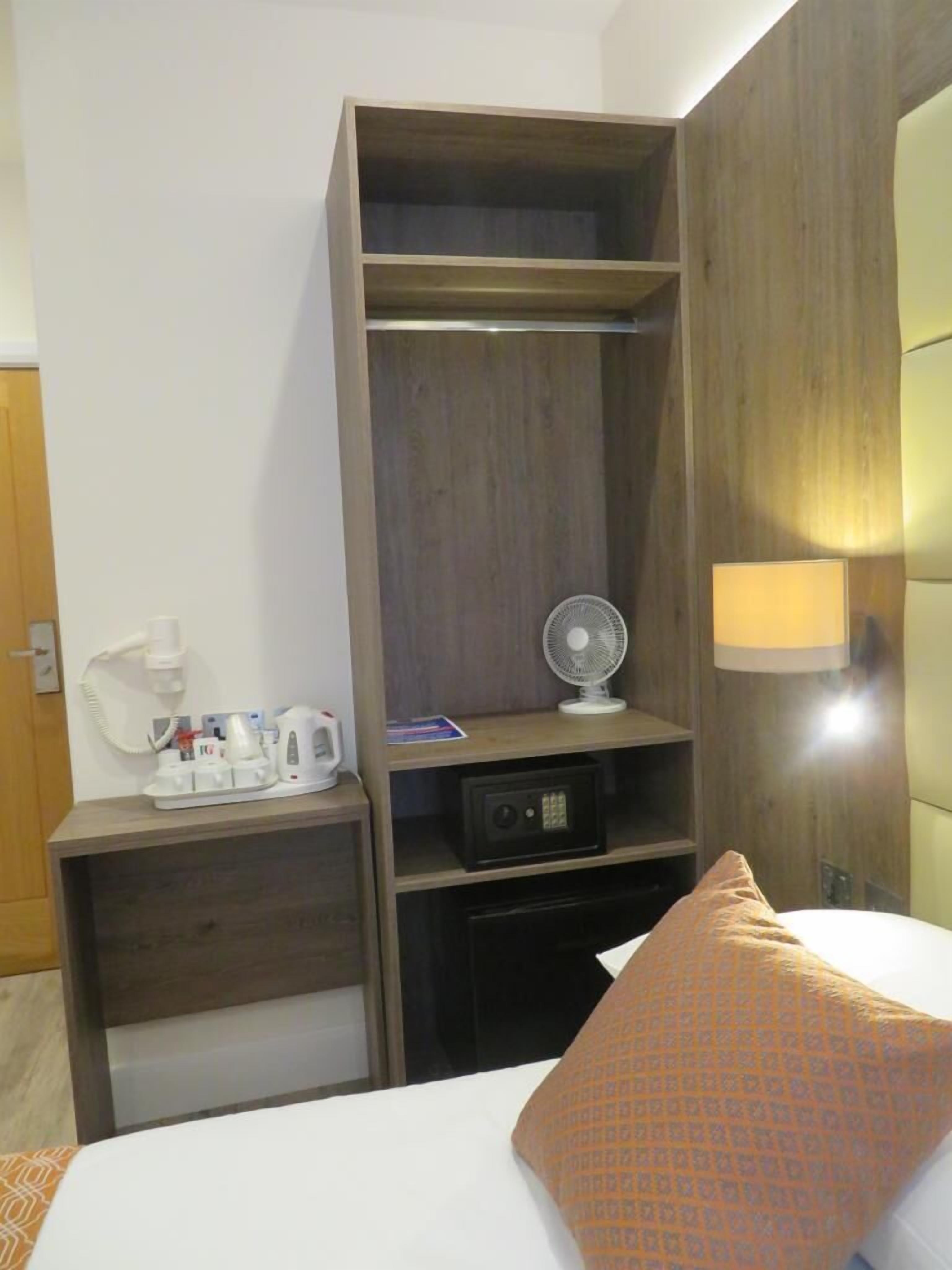 Une chambre d’hôtel comprenant un lit, une table de chevet avec une lampe, un ventilateur et une étagère avec une bouilloire et un téléphone.