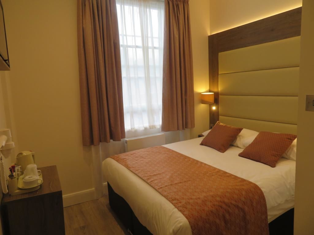 Une chambre d’hôtel avec un grand lit, une lampe de chevet, une tête de lit en bois et une fenêtre avec des rideaux.
