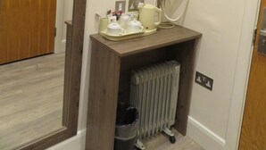 Une chambre d’hôtel avec une porte en bois, un miroir, une petite table avec une bouilloire, des tasses et un téléphone. Un radiateur se trouve sous la table, et un téléviseur est fixé au mur.
