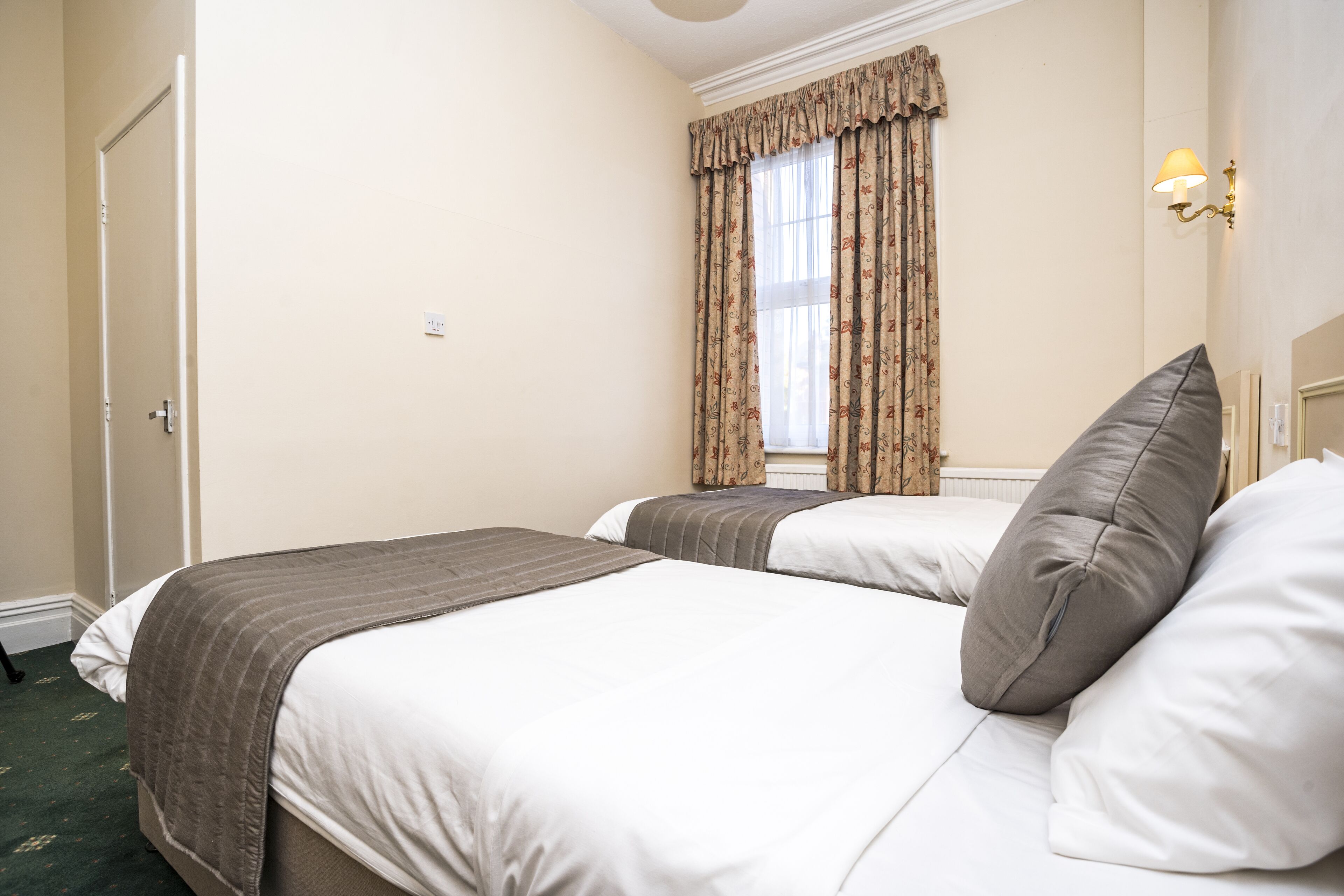 A hotel room with two beds, a window with curtains, a wall-mounted lamp, and a door.
