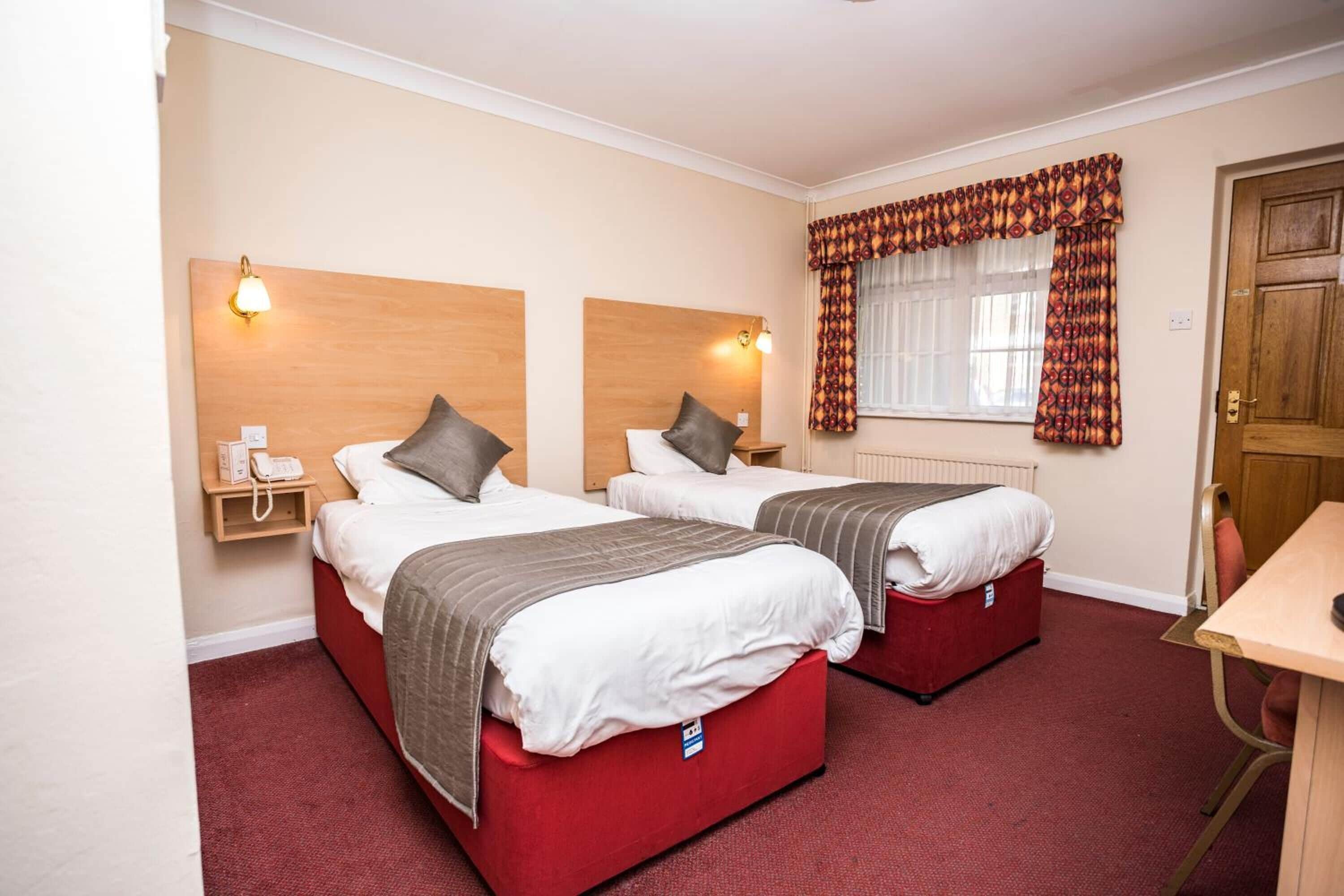 A hotel room with two beds, a wooden headboard, bedside lamps, a telephone, and a window with patterned curtains.