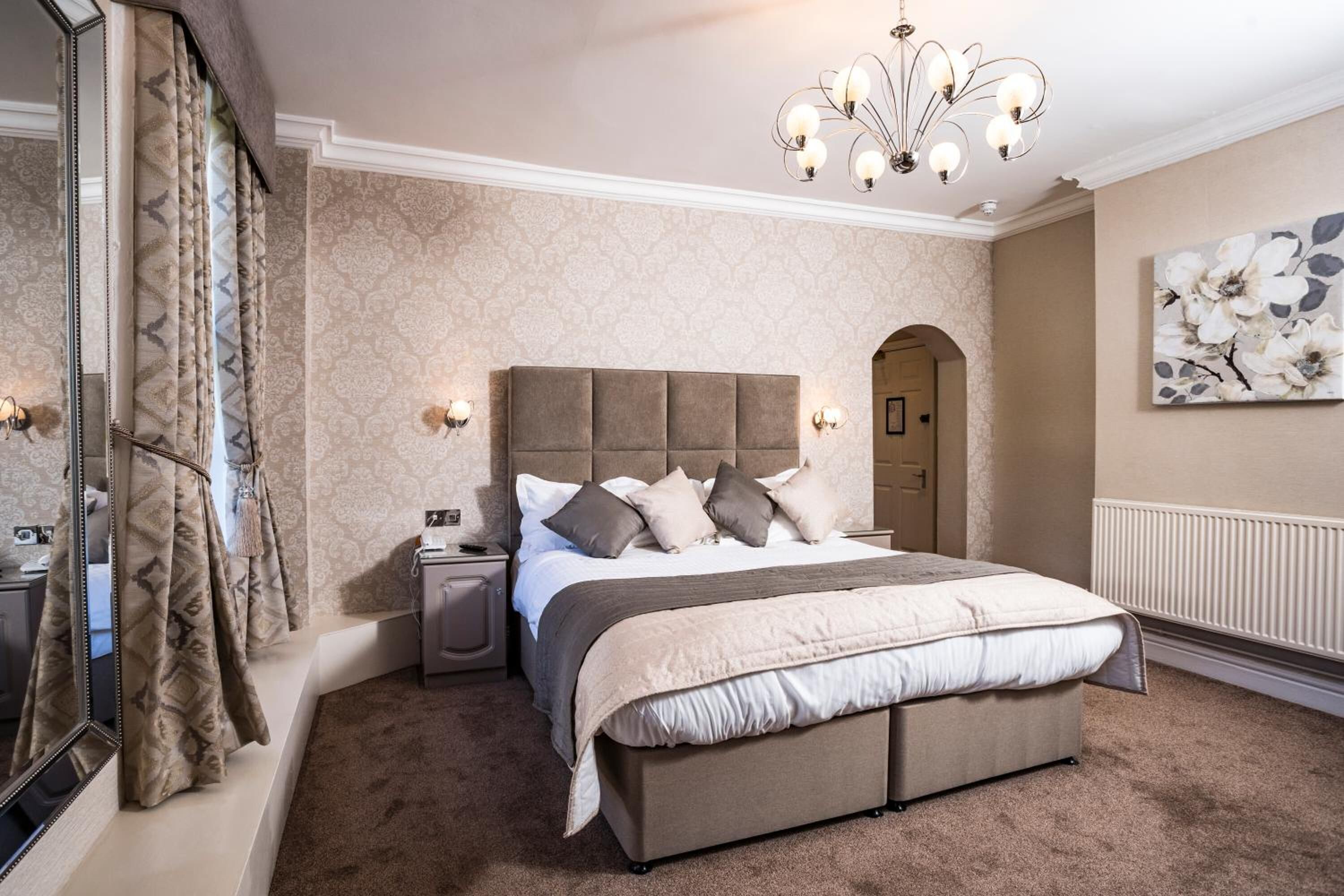 A bedroom with a large bed, a nightstand, a dresser, a mirror, a chandelier, and a door leading to another room.