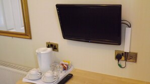 A hotel room with a flat-screen TV mounted on the wall, a coffee maker, and a tray with cups and condiments. - The Fairway Hotel (London)