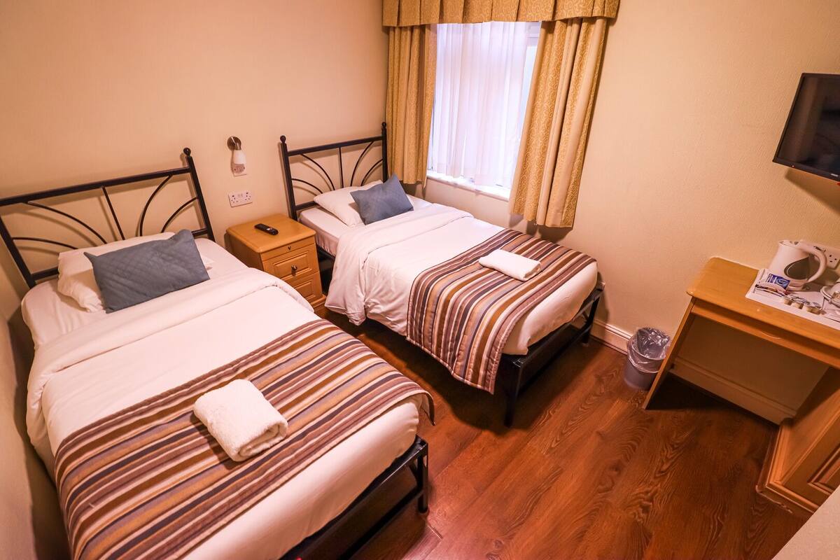 A hotel room with two single beds, a wooden nightstand with a lamp, a small table with a coffee maker, and a window with curtains.