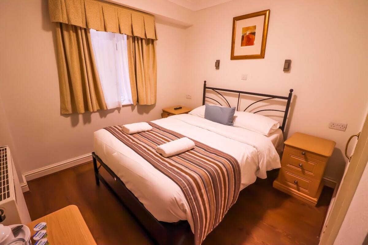 A neatly made bed with striped bedding and two pillows, a bedside table with a lamp, a small wooden nightstand, and a window with curtains.