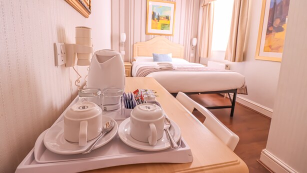 A hotel room with a neatly arranged breakfast tray, a bed with a headboard, a bedside table, and a window with curtains. - The Fairway Hotel (London)