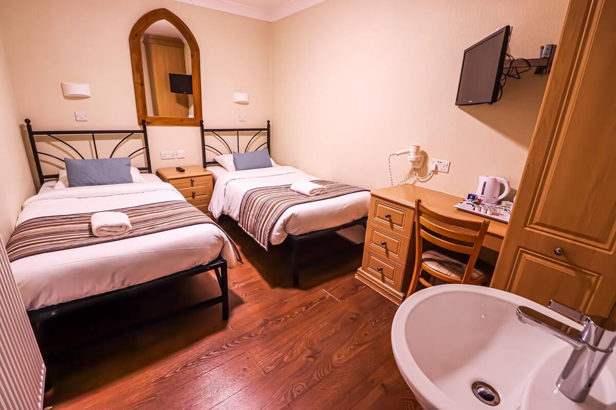 A hotel room with two beds, a wooden dresser, a desk with a chair, a TV mounted on the wall, and a bathroom with a sink and mirror.