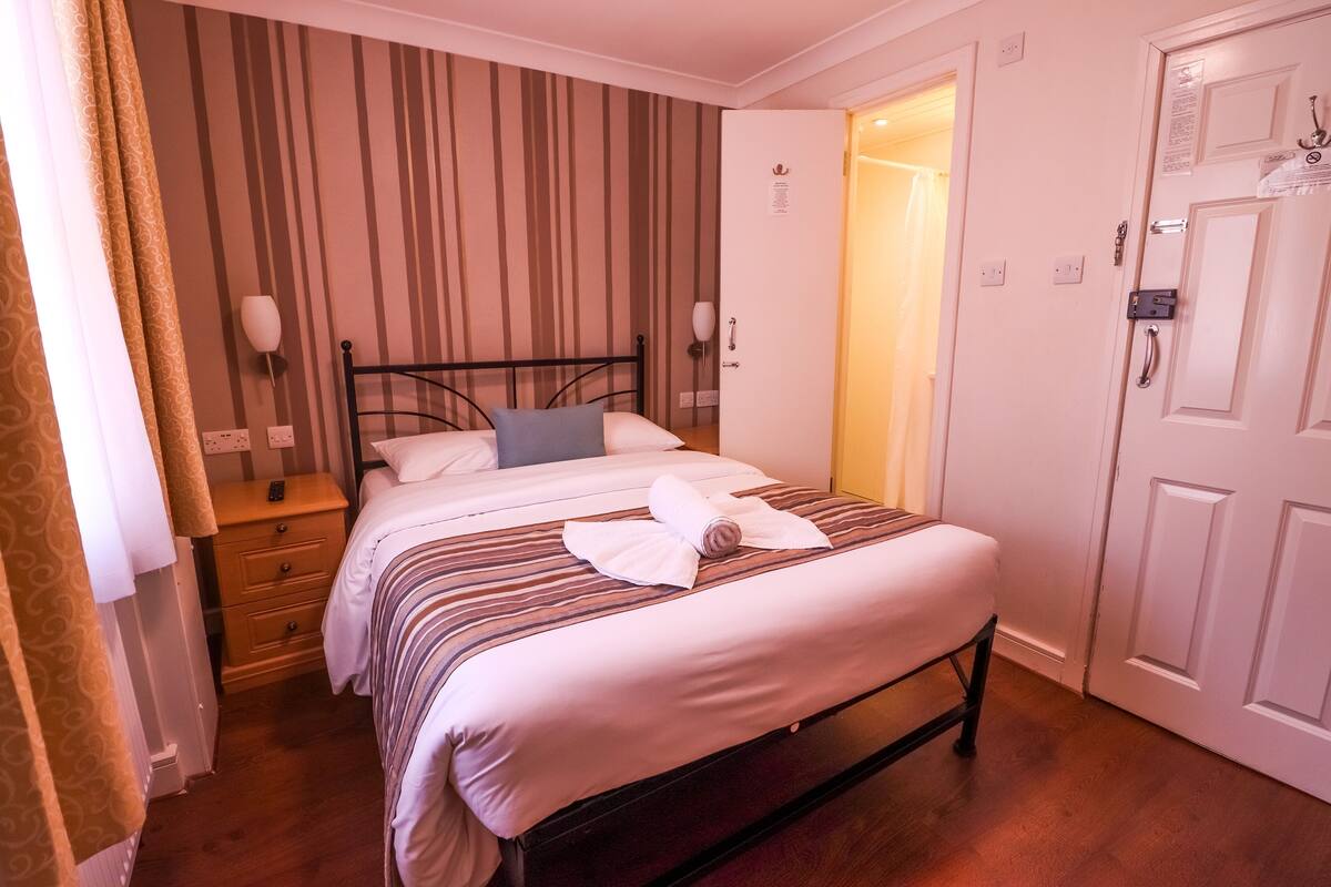 A neatly made bed with a rolled towel, a bedside table with a lamp, and a door with a lock.