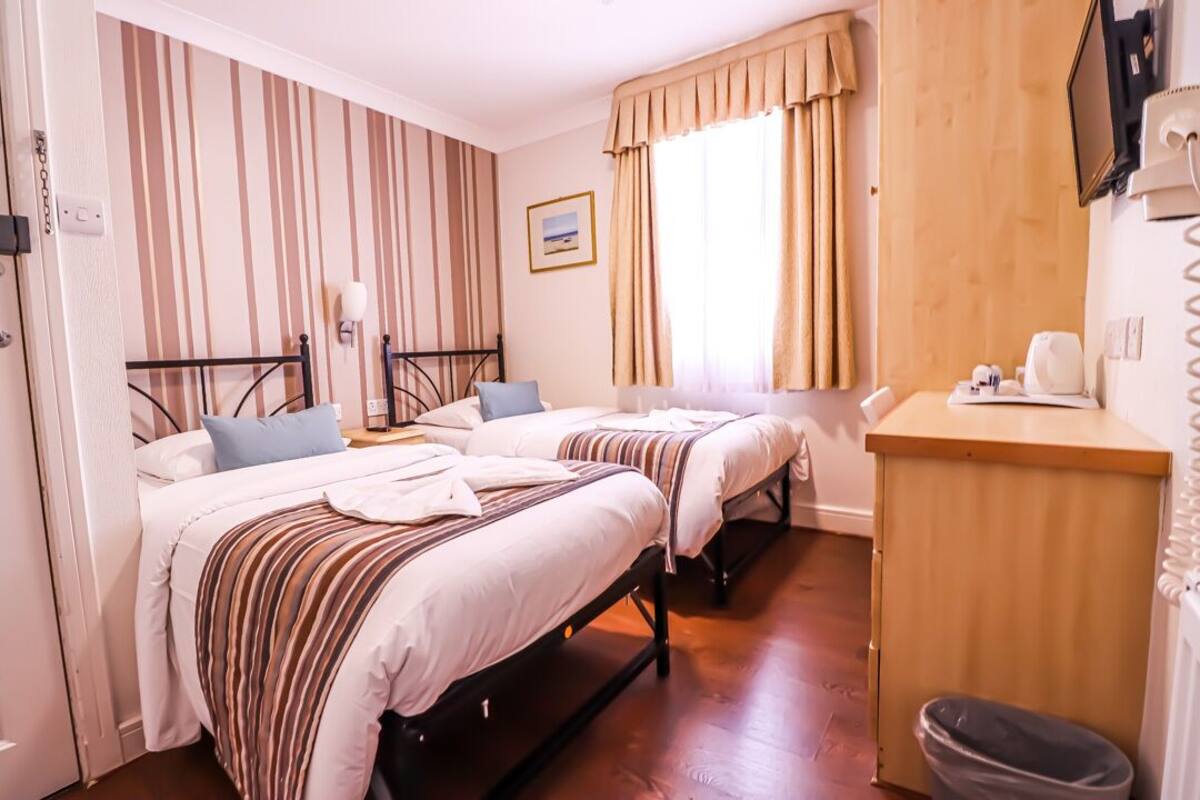 A hotel room with two beds, a wooden desk, a trash bin, and a window with curtains.