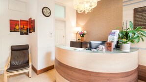 A reception desk with a clock, framed pictures, a chair, a lamp, and a sign indicating free Wi-Fi. - The Fairway Hotel (London)