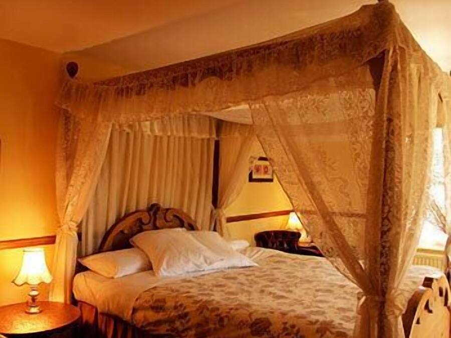 A four-poster bed with a canopy, a bedside table with a lamp, and a chair in a warmly lit room.