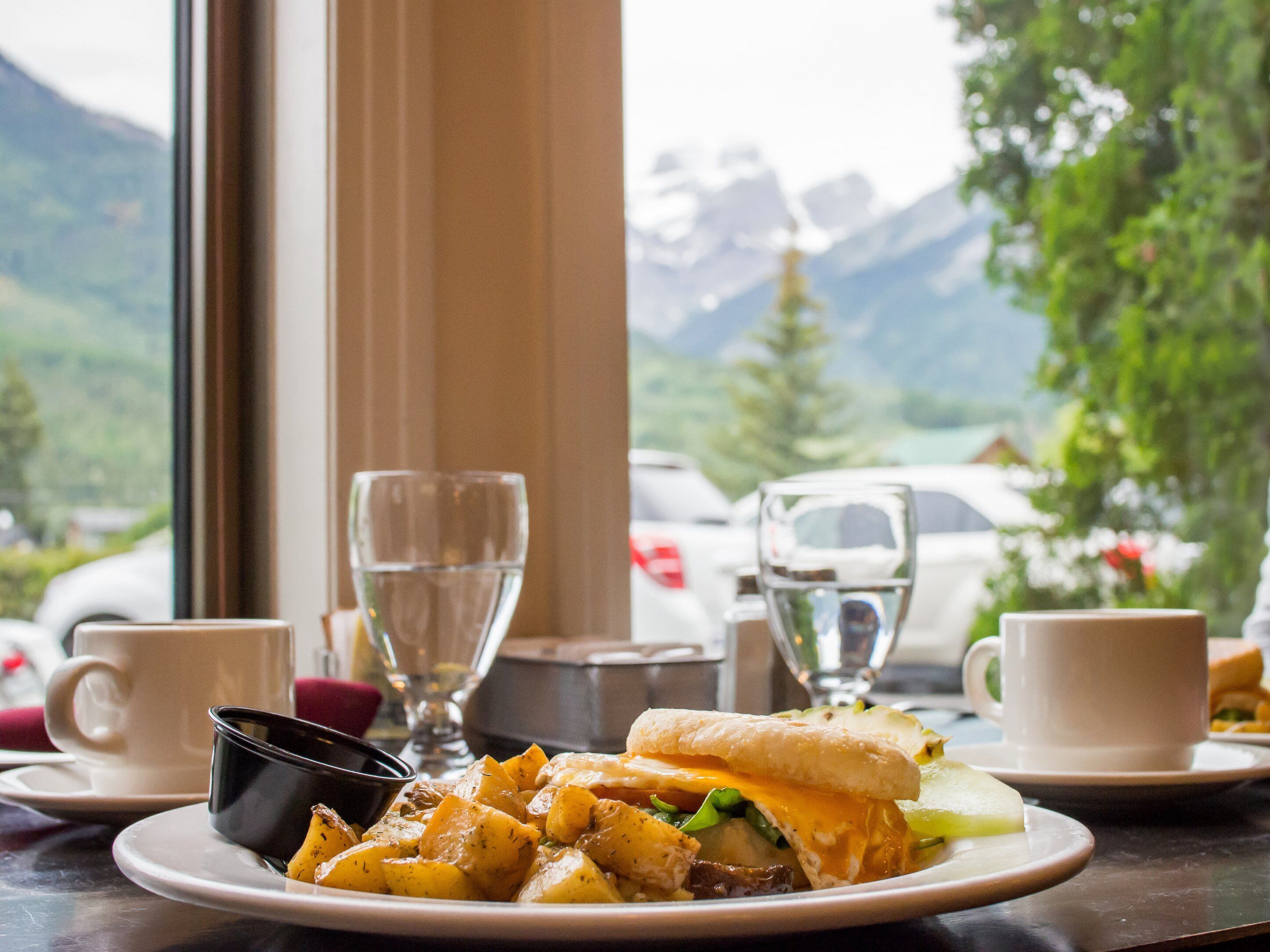 A meal with a sandwich and potatoes, two cups, and a glass on a table with a mountain view in the background.