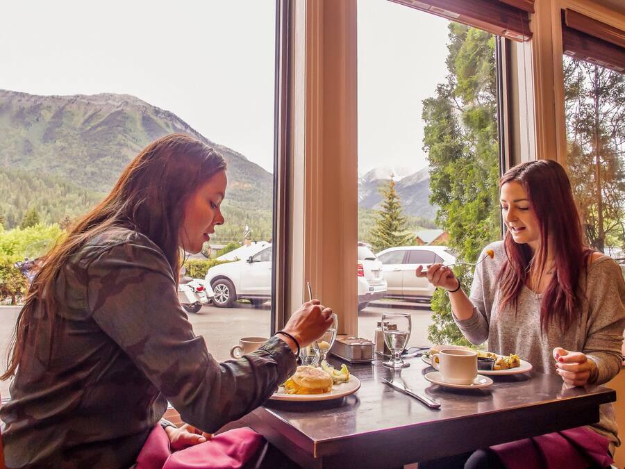 Dos personas sentadas en una mesa con vistas a la montaña, un coche estacionado afuera y una ventana que da a árboles y montañas.