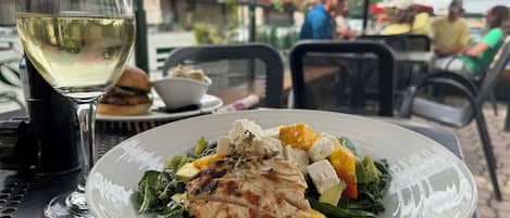 A meal with grilled chicken and vegetables on a white plate, a glass of white wine, and a table with a metal grid pattern.