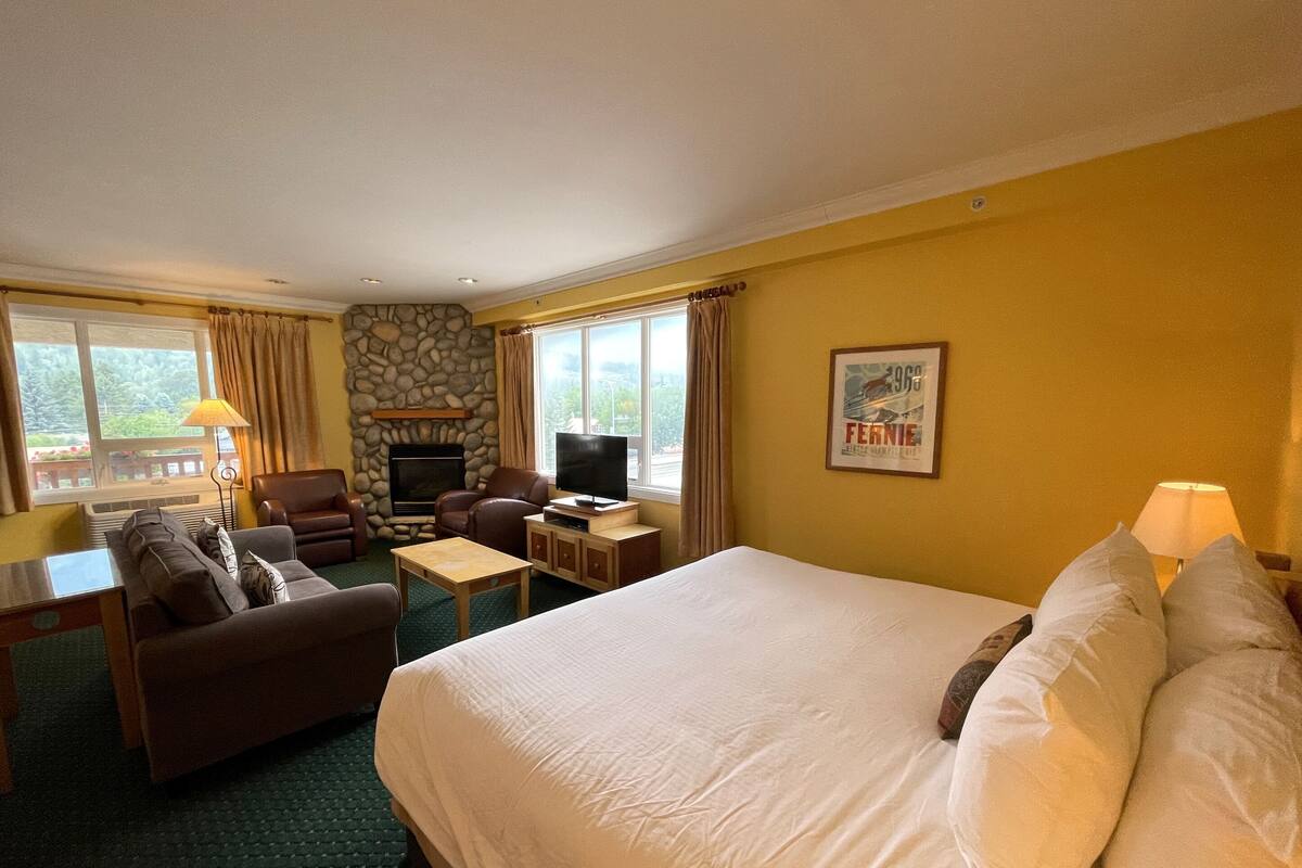 Una habitación de hotel con una cama grande, un sofá, un televisor, una chimenea y un ventanal con cortinas.