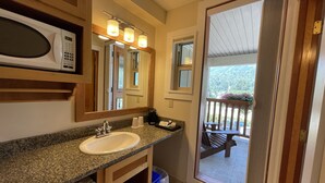 A bathroom with a granite countertop, a white sink, a mirror, a microwave, and a view of a balcony with outdoor furniture. - Park Place Lodge (Fernie)