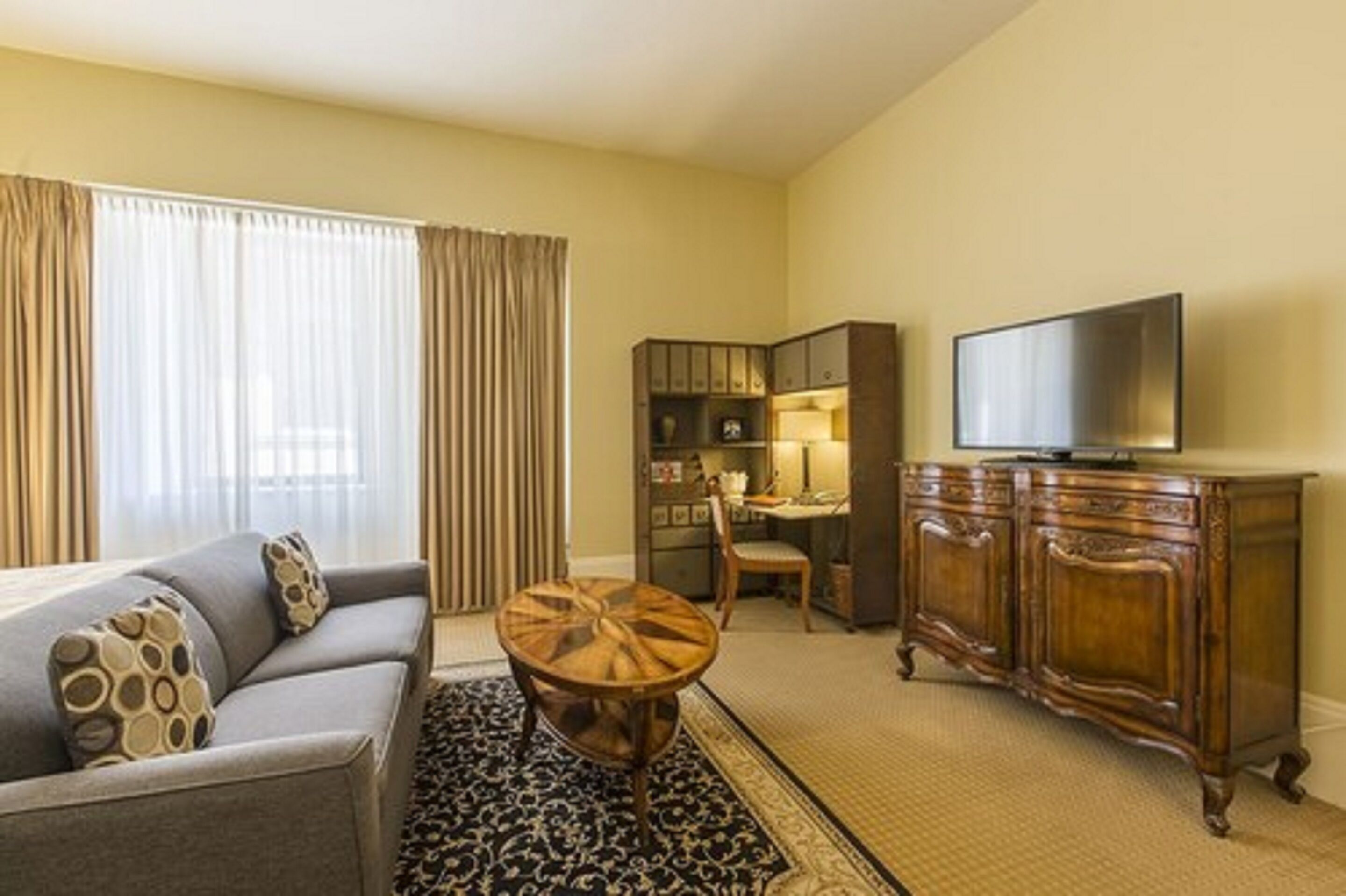 A hotel room with a sofa, a round wooden table, a TV on a wooden cabinet, and a desk with a chair.
