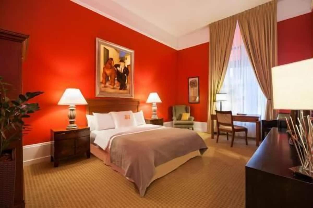 A hotel room with a large bed, two bedside tables with lamps, a desk with a chair, and a painting on the wall.
