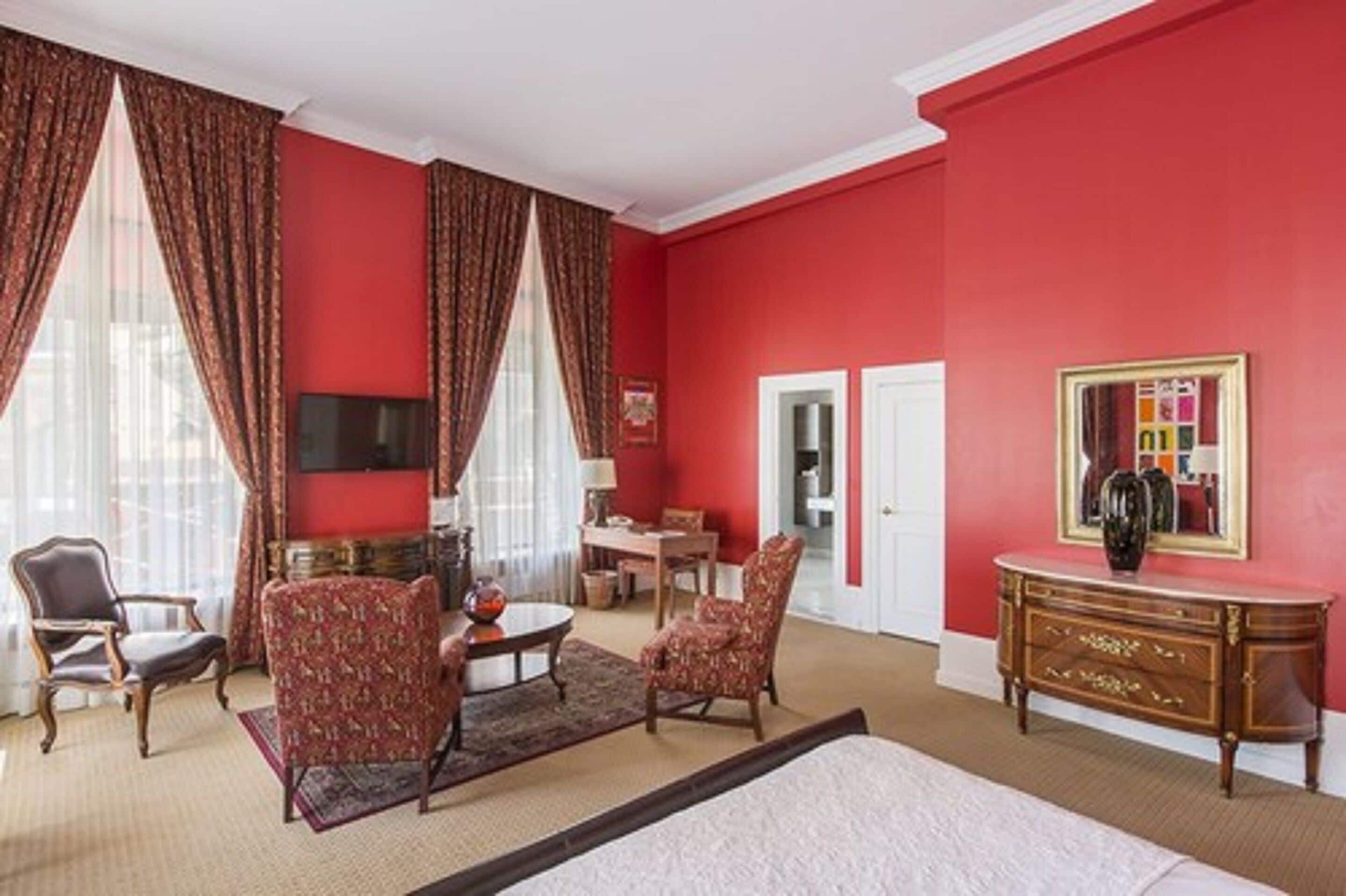 A hotel room with a red wall, a bed, a TV, a desk, and a chair.