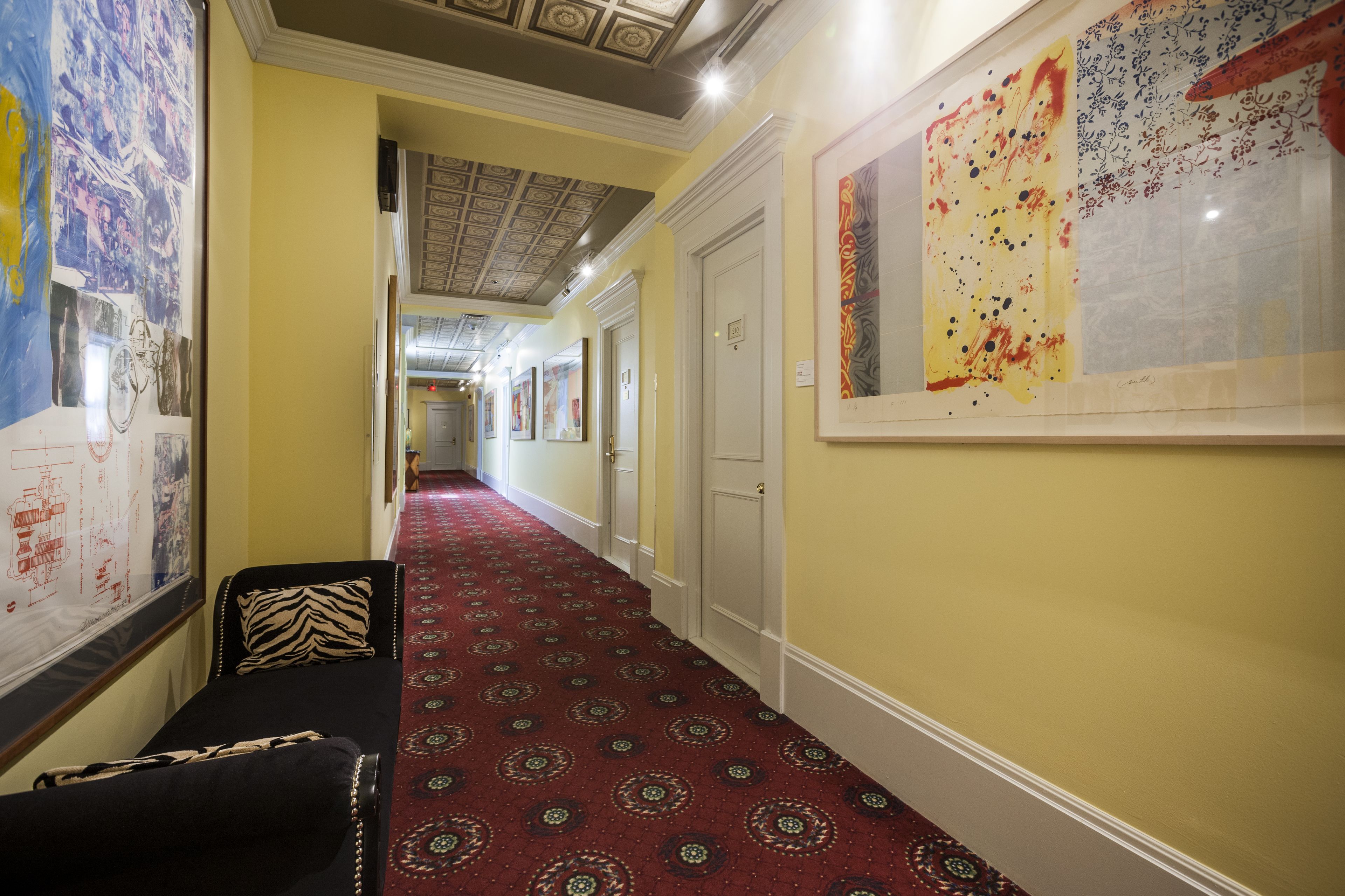 A hotel corridor with patterned carpet, a zebra-patterned pillow, and artwork on the walls.